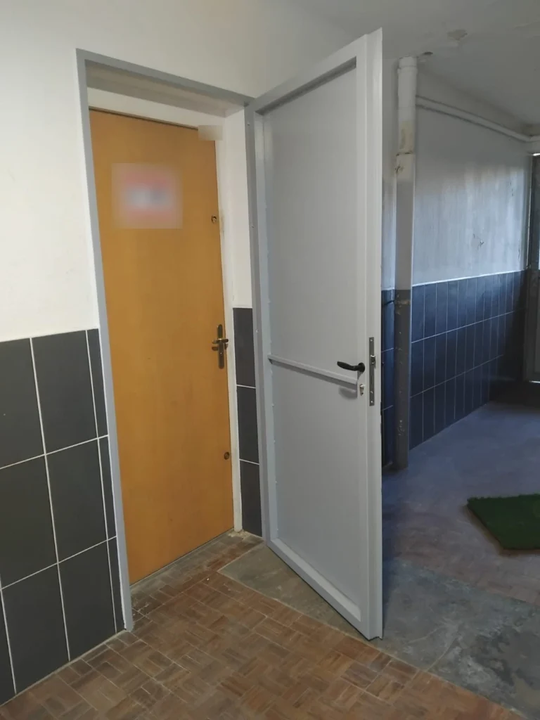 Porte en acier gris ouverte, posée devant une porte d’entrée en bois